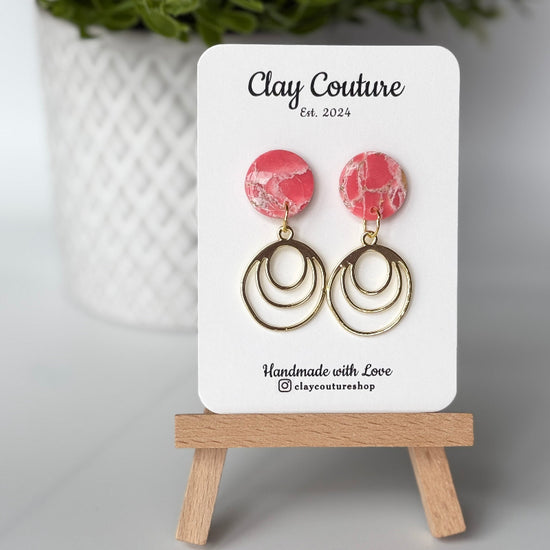 Polymer clay marbled earrings in blush color with resin finish and brass charms on a 'Clay Couture' branded card with a plant in the background.