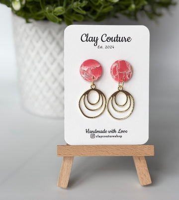 Polymer clay marbled earrings in blush color with resin finish and brass charms on a 'Clay Couture' branded card with a plant in the background.