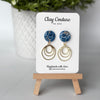 Polymer clay marbled earrings in Midnight blue  color with resin finish and brass charms on a 'Clay Couture' branded card with a plant in the background.