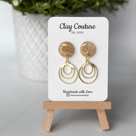 Polymer clay marbled earrings in warm sandstone color with resin finish and brass charms on a 'Clay Couture' branded card with a plant in the background.