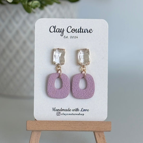 Mauvepolymer clay earrings with a crystal stud on a 'Clay Couture' branded card with a wooden stand.