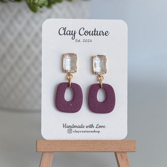 Plum polymer clay earrings with a crystal stud on a 'Clay Couture' branded card with a wooden stand.