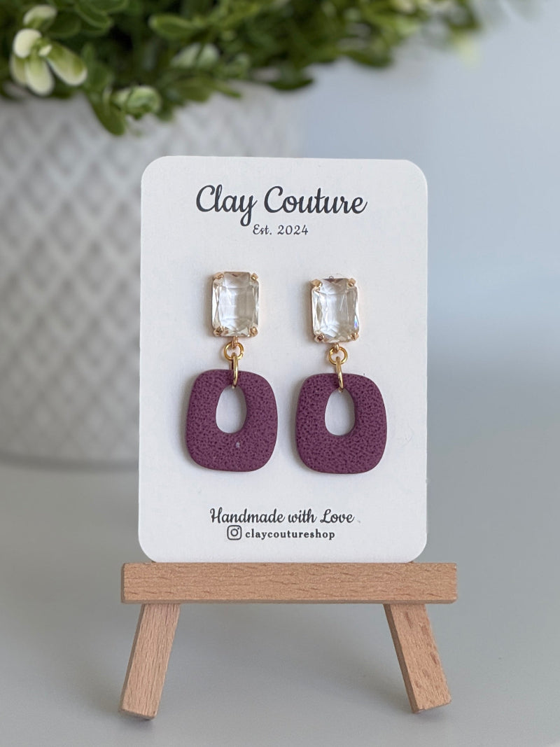 Plum polymer clay earrings with a crystal stud on a 'Clay Couture' branded card with a wooden stand.