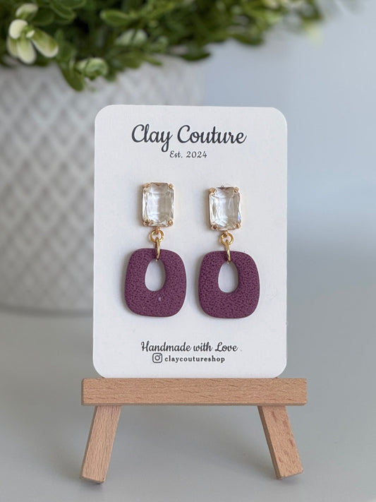 Plum polymer clay earrings with a crystal stud on a 'Clay Couture' branded card with a wooden stand.