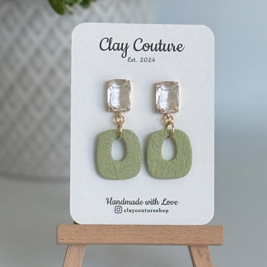 Sage polymer clay earrings with a crystal stud on a 'Clay Couture' branded card with a wooden stand.