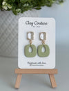Sage polymer clay earrings with a crystal stud on a 'Clay Couture' branded card with a wooden stand.