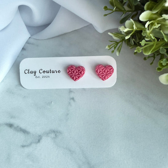Pink heart-shaped earrings on a Clay Couture branded card with greenery in the background.