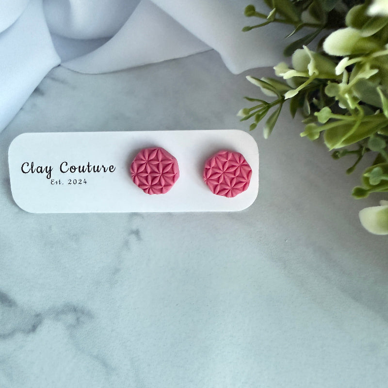 Pink geometric earrings on a white card with 'Clay Couture' branding, placed on a marble surface with greenery.