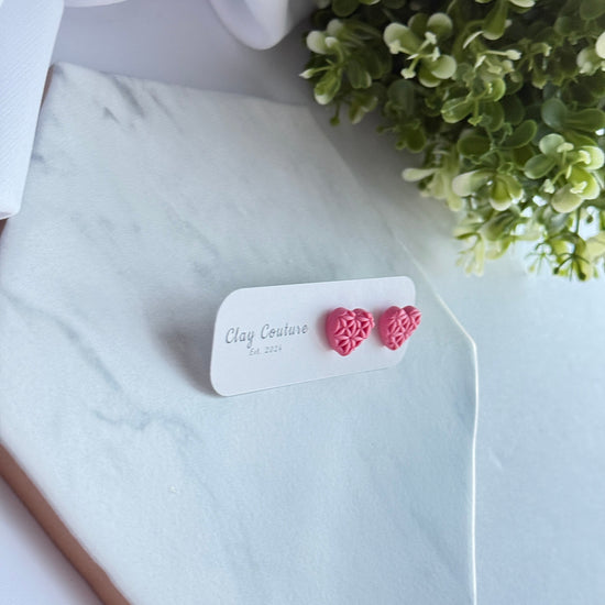 Pink heart-shaped earrings on a white card with 'Clay Couture' branding, placed on a marble surface with greenery.