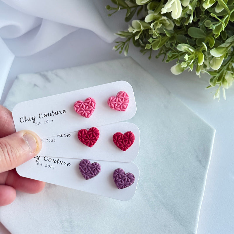 Set of heart-shaped earrings on a Clay Couture card with a white background and greenery.