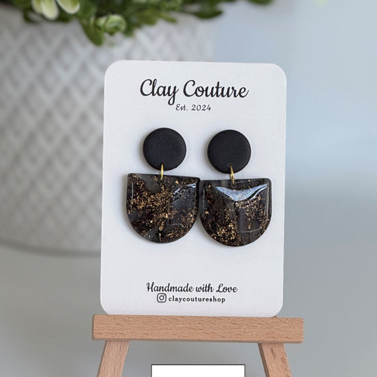 Black circular earrings on a 'Clay Couture' branded card with a wooden stand.