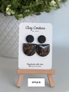 Black circular earrings on a 'Clay Couture' branded card with a wooden stand.
