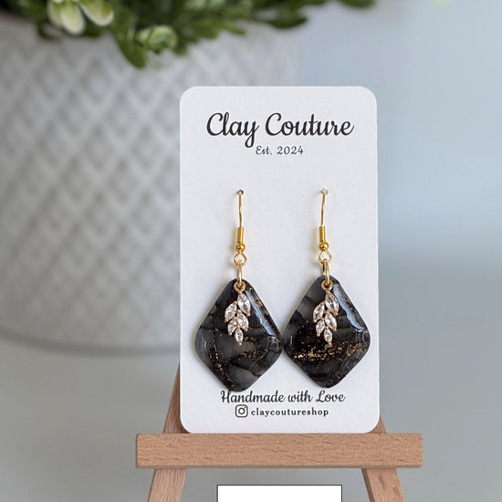 Black  and gold diamond-shaped earrings on a 'Clay Couture' branded card with a wooden stand.