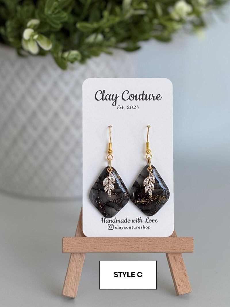 Black  and gold diamond-shaped earrings on a 'Clay Couture' branded card with a wooden stand.