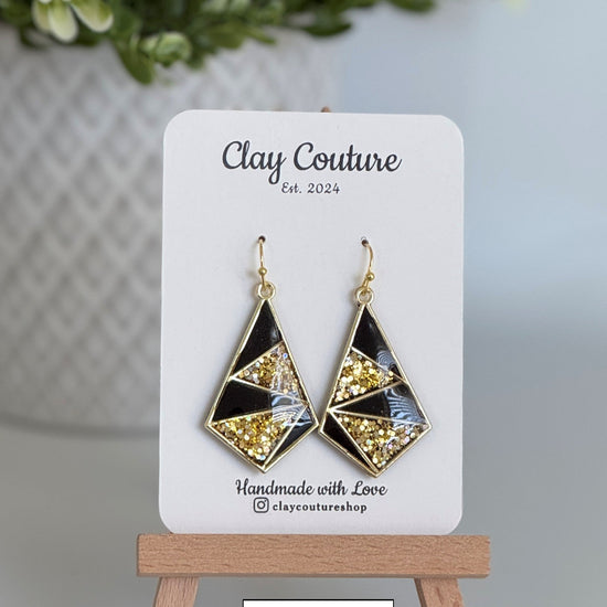 Geometric earrings on a Clay Couture display card with a blurred plant in the background.