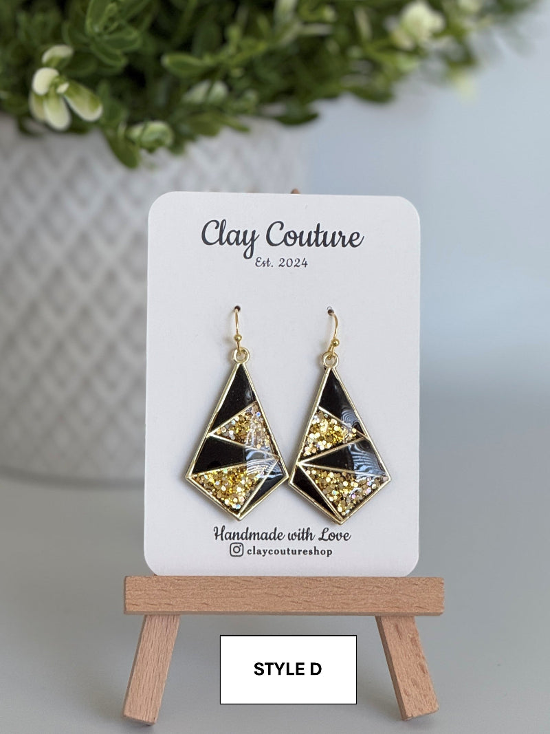 Geometric earrings on a Clay Couture display card with a blurred plant in the background.