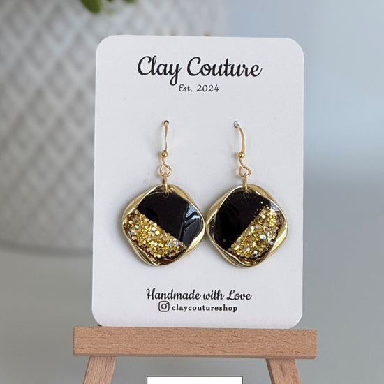 Black and gold earrings on a 'Clay Couture' branded card with a wooden stand.