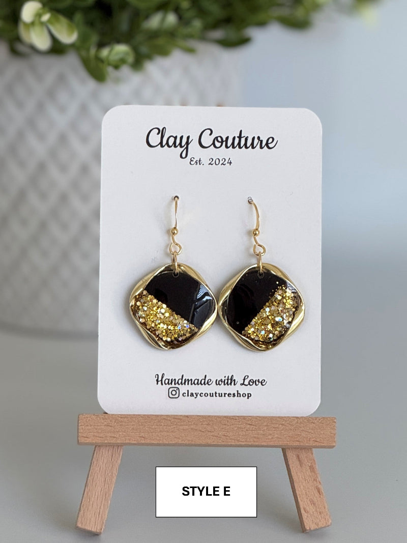 Black and gold earrings on a 'Clay Couture' branded card with a wooden stand.