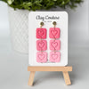 Pink heart-shaped earrings on a 'Clay Couture' branded card with a wooden stand.