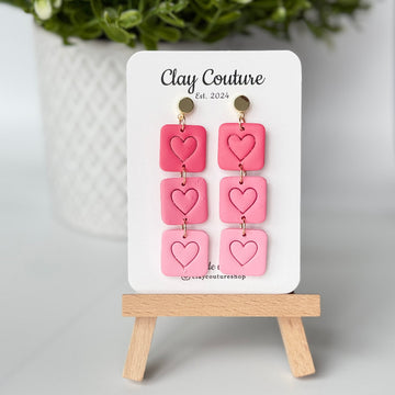 Pink heart-shaped earrings on a 'Clay Couture' branded card with a wooden stand.