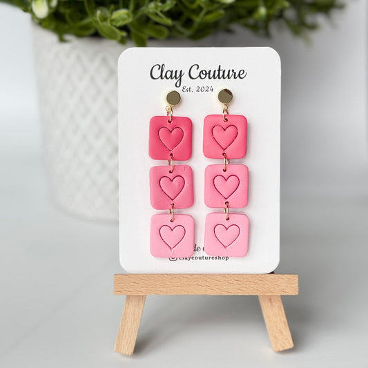 Pink heart-shaped earrings on a 'Clay Couture' branded card with a wooden stand.