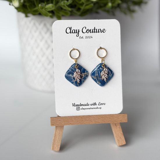 Pair of marbled polymer clay earrings with resin finish  in midnight blue with crystal charm and gold plated studs on Clay Couture branded card on a wooden stand