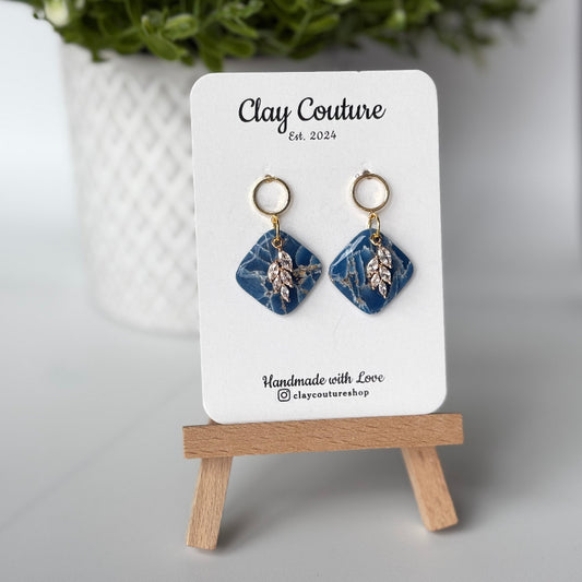 Pair of marbled polymer clay earrings with resin finish  in midnight blue with crystal charm and gold plated studs on Clay Couture branded card on a wooden stand