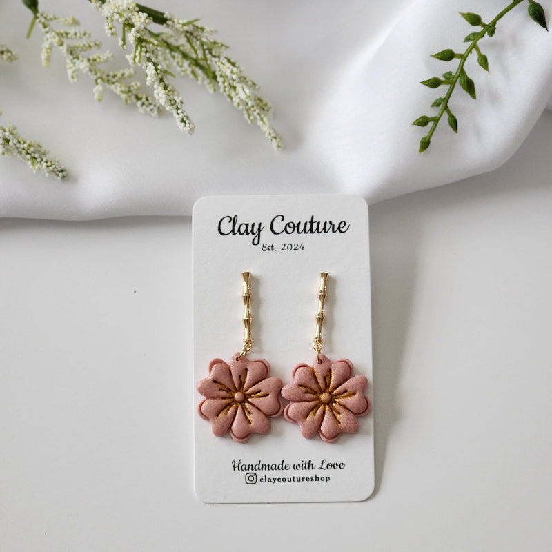 Floral earrings in rose pink color on a Clay Couture branded card with white flowers and green leaves in the background.