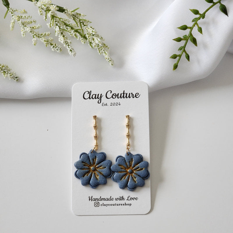 Blue flower earrings on a Clay Couture branded card with white flowers and green leaves in the background.