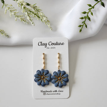 Blue flower earrings on a Clay Couture branded card with white flowers and green leaves in the background.