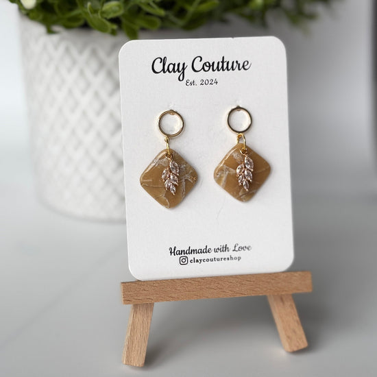 Pair of marbled polymer clay earrings with resin finish  in warm sandstone with crystal charm and gold plated studs on Clay Couture branded card on a wooden stand