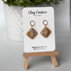 Pair of marbled polymer clay earrings with resin finish  in warm sandstone with crystal charm and gold plated studs on Clay Couture branded card on a wooden stand