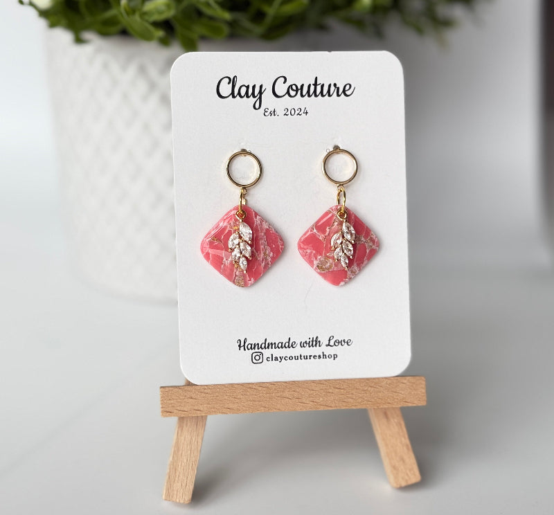 Pair of marbled polymer clay earrings with resin finish  in blush with crystal charm and gold plated studs on Clay Couture branded card on a wooden stand