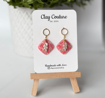 Pair of marbled polymer clay earrings with resin finish  in blush with crystal charm and gold plated studs on Clay Couture branded card on a wooden stand