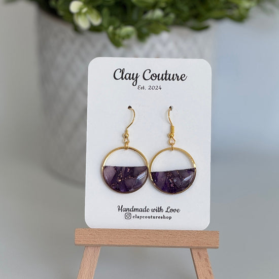 Amethyst earrings on a 'Clay Couture' branded card with a plant in the background.