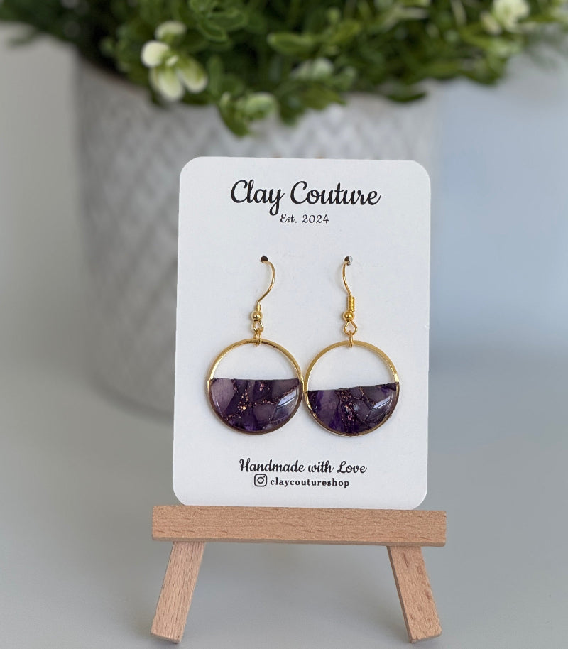 Amethyst earrings on a 'Clay Couture' branded card with a plant in the background.