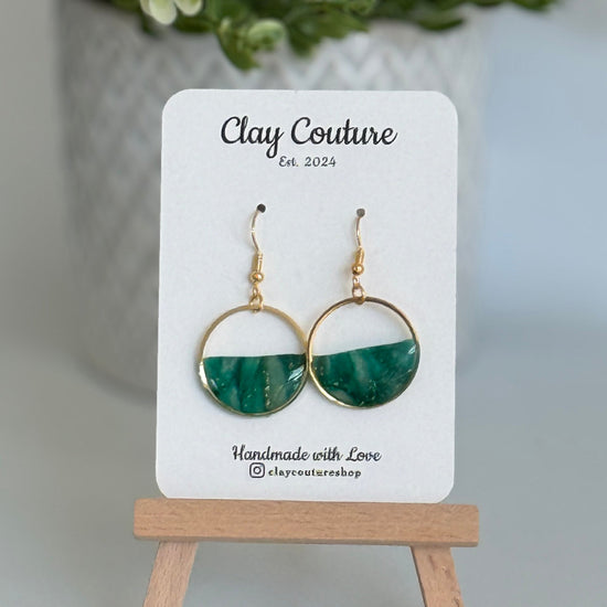 Green and gold hoop earrings on a 'Clay Couture' branded card with a plant in the background.