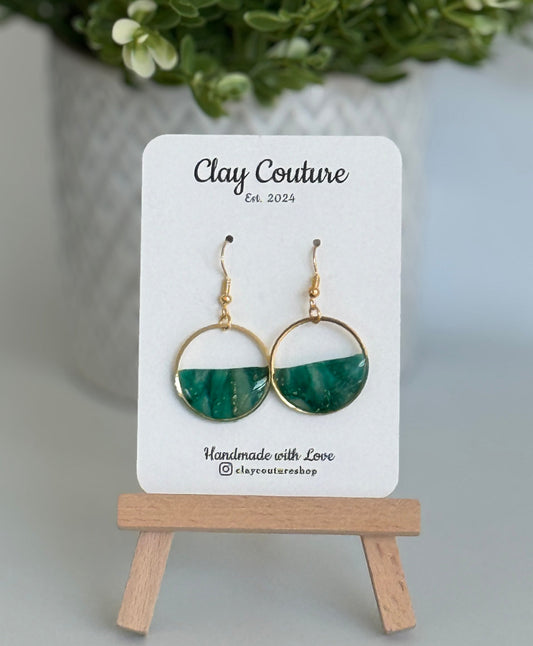 Green and gold hoop earrings on a 'Clay Couture' branded card with a plant in the background.