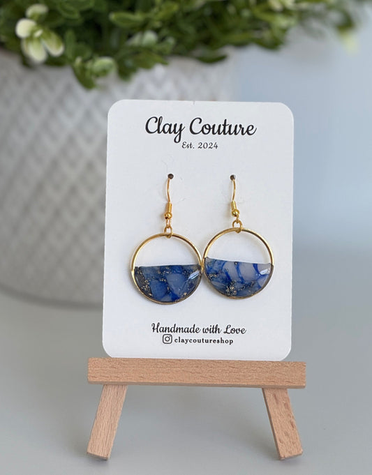 Pair of blue and gold earrings on a 'Clay Couture' branded card with a wooden stand.