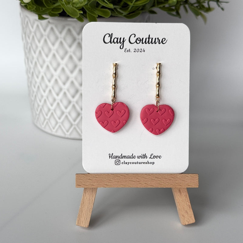 Heart-shaped earrings in Blush Rose color on a 'Clay Couture' branded card with a wooden stand.
