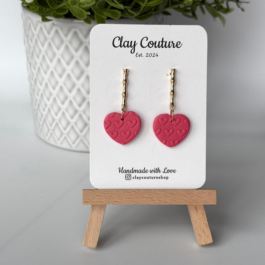 Heart-shaped earrings in Blush Rose color on a 'Clay Couture' branded card with a wooden stand.