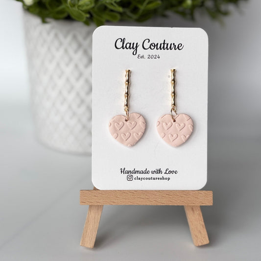 Heart-shaped earrings in champagne color on a 'Clay Couture' branded card with a wooden stand.