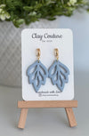 Embossed leaf design  earrings in Duty Blue color with gold plated hoops on a 'Clay Couture' branded card with a plant in the background.