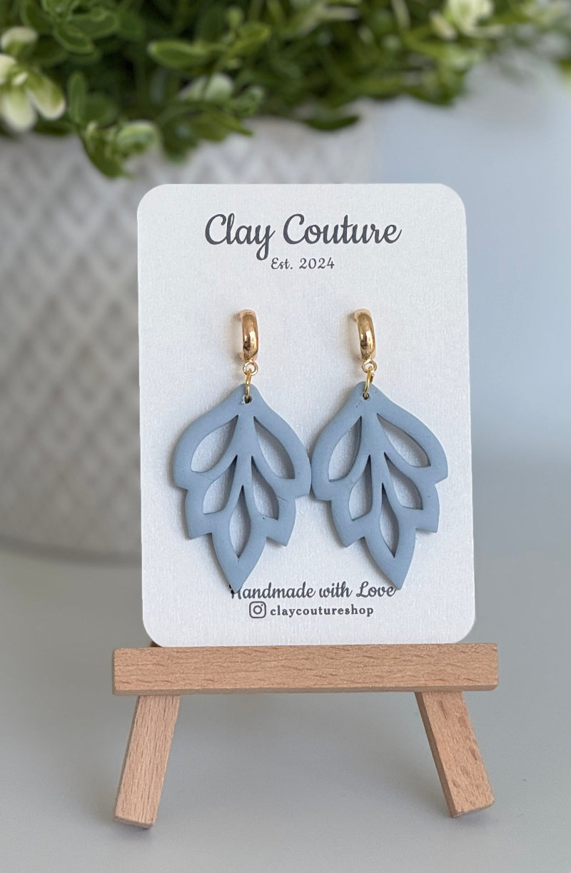 Embossed leaf design  earrings in Duty Blue color with gold plated hoops on a 'Clay Couture' branded card with a plant in the background.