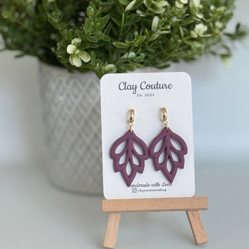 Embossed leaf design  earrings in Plum color with gold plated hoops on a 'Clay Couture' branded card with a plant in the background.