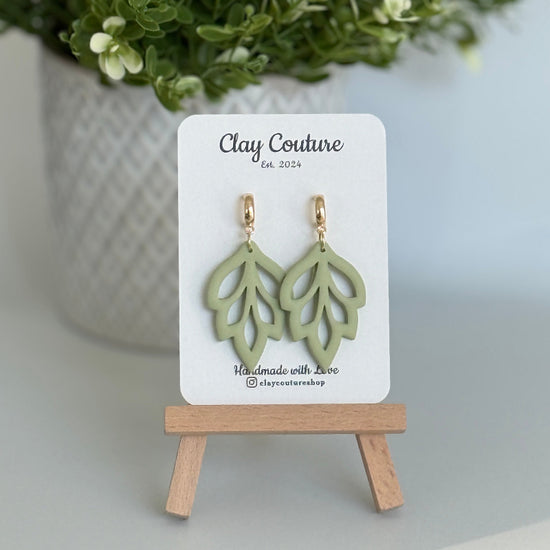 Embossed leaf design  earrings in Sage color with gold plated hoops on a 'Clay Couture' branded card with a plant in the background.