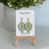 Embossed leaf design  earrings in Sage color with gold plated hoops on a 'Clay Couture' branded card with a plant in the background.