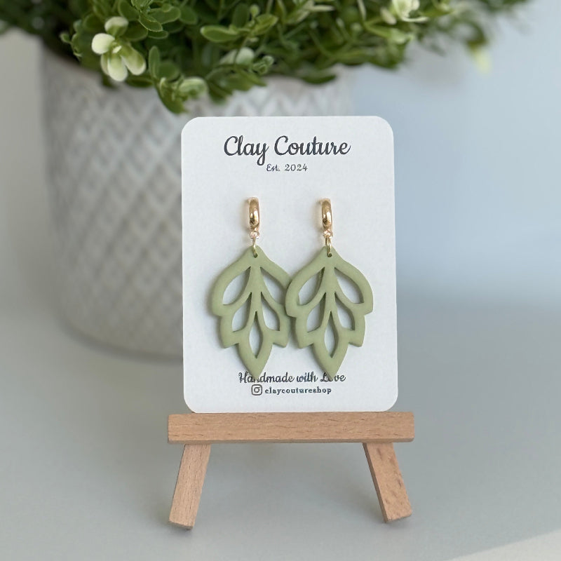 Embossed leaf design  earrings in Sage color with gold plated hoops on a 'Clay Couture' branded card with a plant in the background.