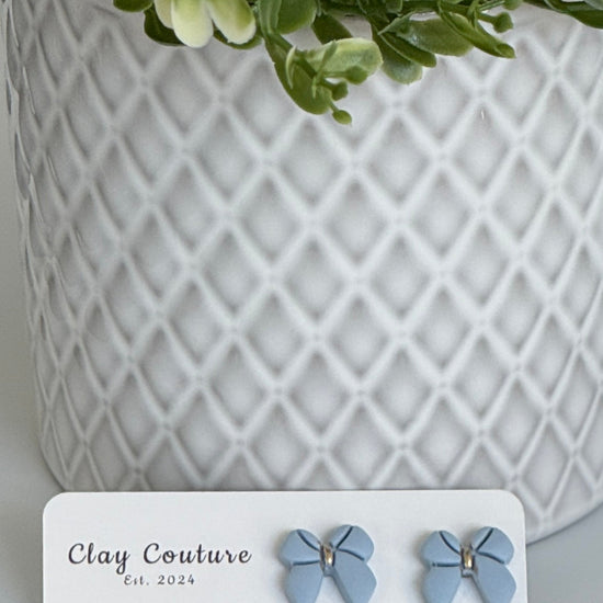 Dusty Blue color Polymer Clay bow earrings on a Clay Couture card with a white textured background