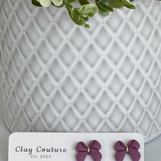Plum color Polymer Clay bow earrings on a Clay Couture card with a white textured background